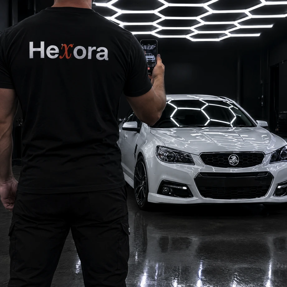 A Hexora videographer, logo on the back of his shirt, filming a white sedan under hexagon lights in a studio.